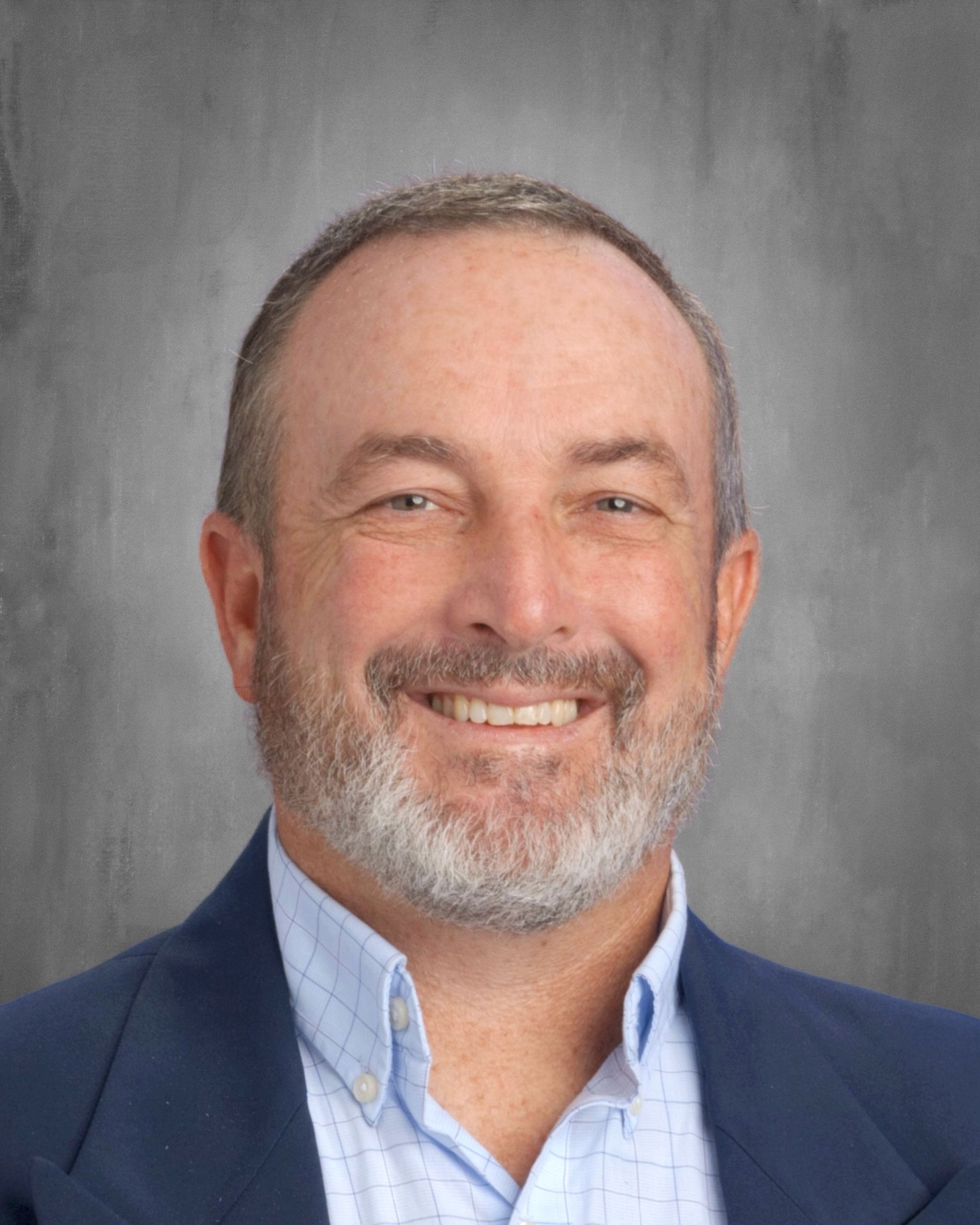 Smiling man with a grey beard, blue blazer, and button-down shirt; gray background.