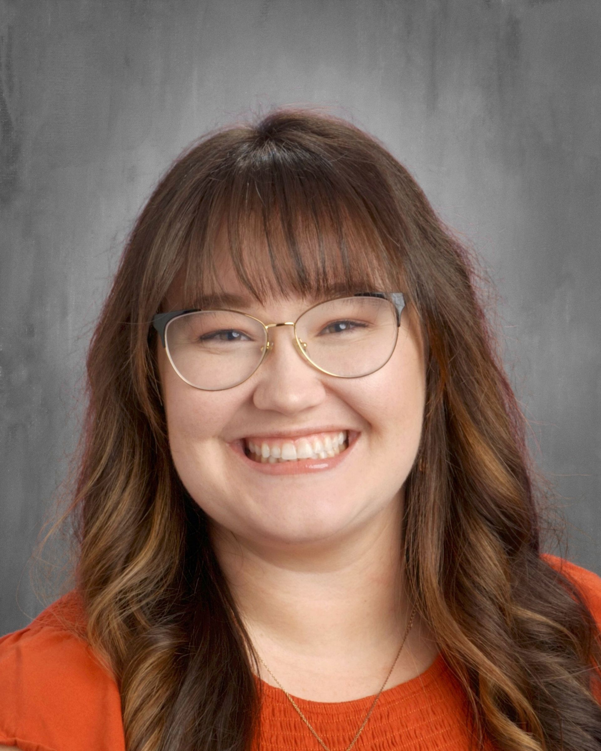 Woman with glasses smiles, wearing an orange top, in front of a gray backdrop.