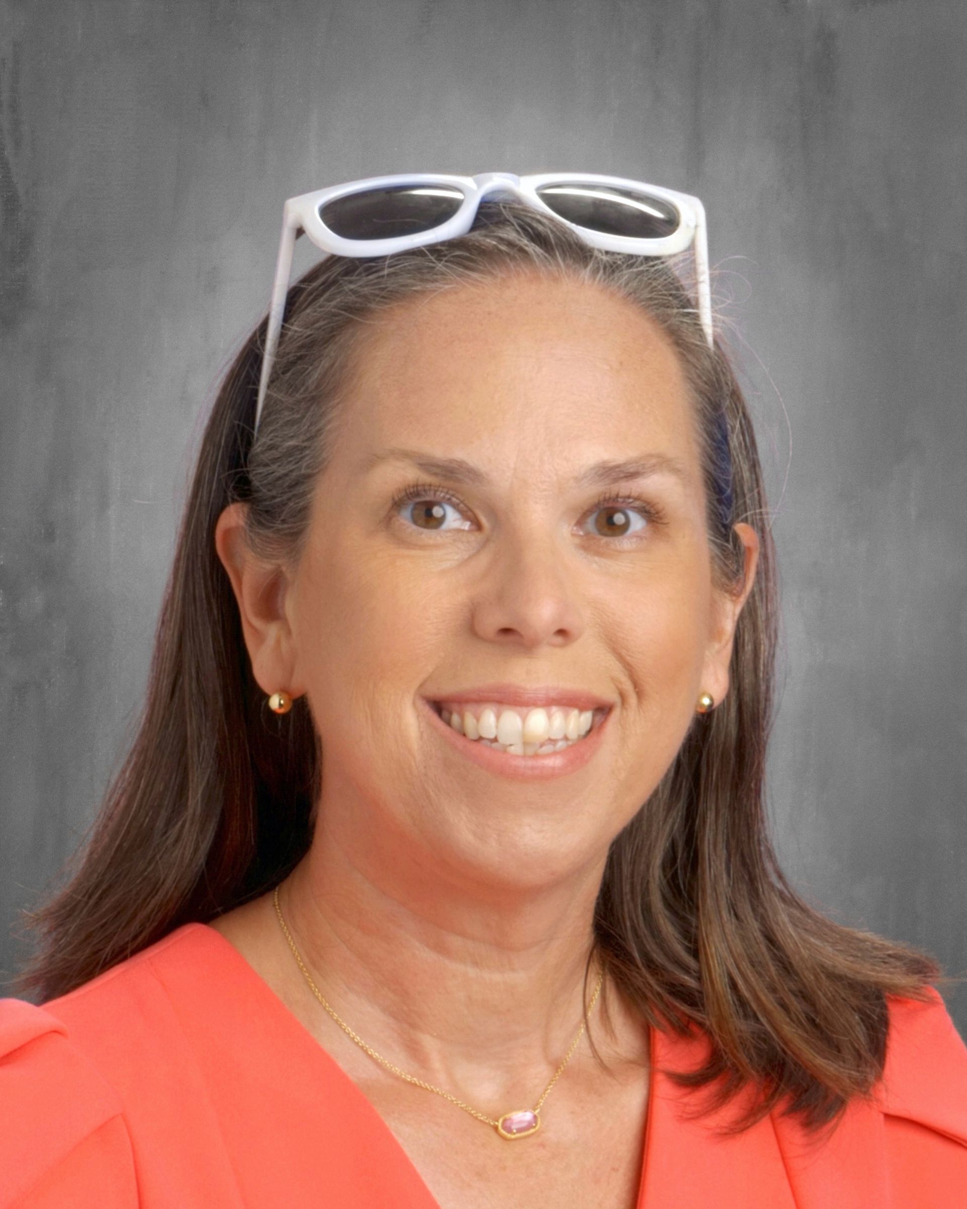 Woman with glasses on head smiles, wearing orange top, gold jewelry, gray background.