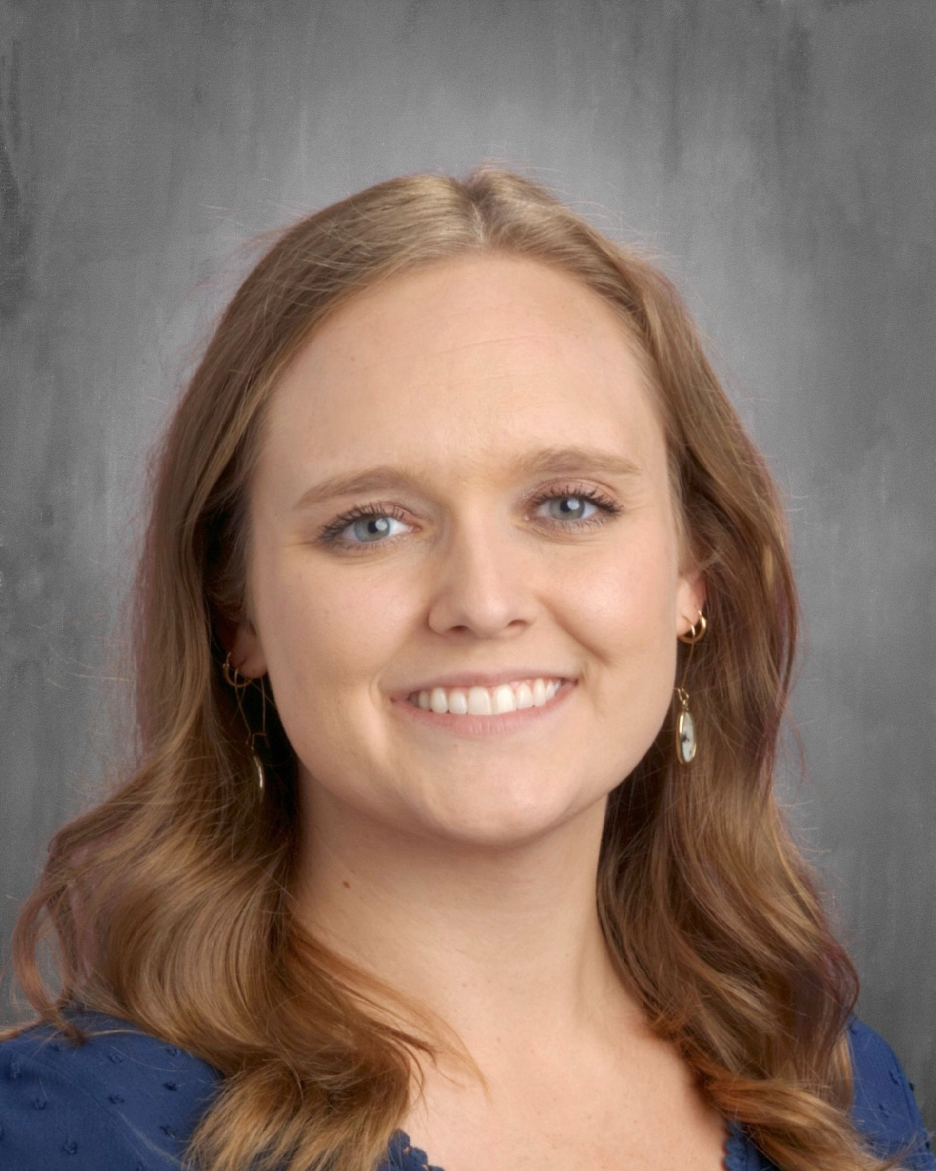Smiling woman with light brown hair and blue eyes. Wearing a dark blue top, posing in front of a gray background.