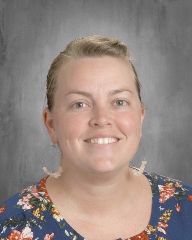 Woman smiling, with fair skin and blue eyes, wearing a floral shirt, standing against a gray background.