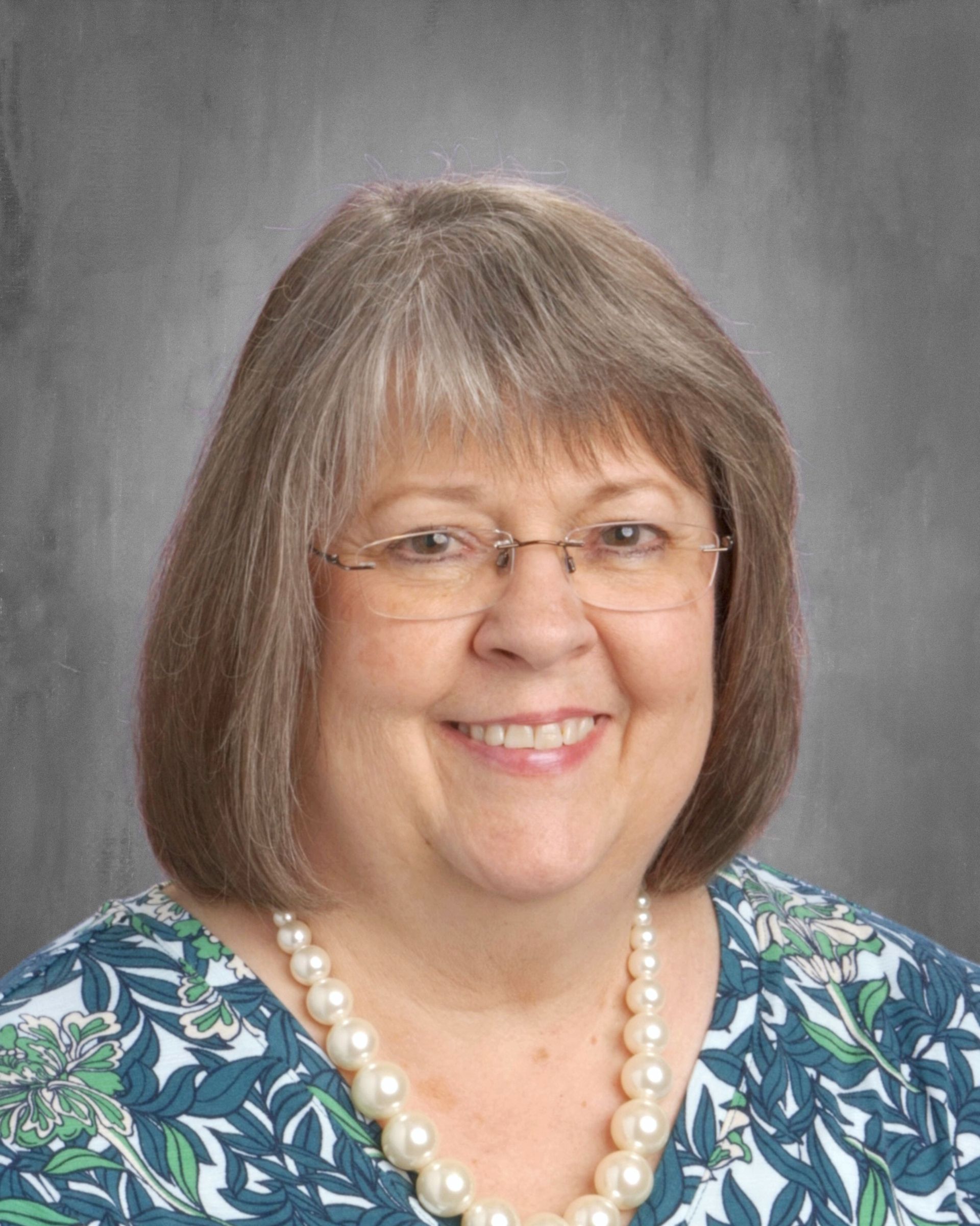 Smiling woman with glasses, short hair, pearl necklace, wearing patterned top.