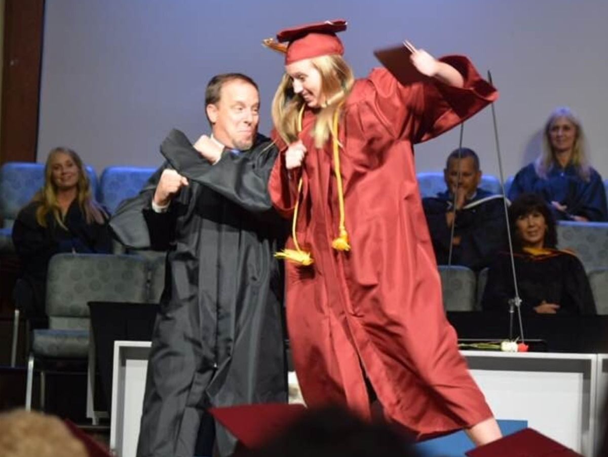 Man in black robe hugs ecstatic graduate in red gown, onstage at graduation.