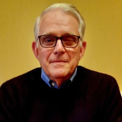 Older man with glasses, wearing a black sweater over a blue shirt, neutral expression, against a yellow background.