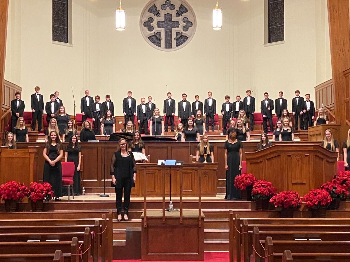 Choir performing on stage in a church. Men in tuxedos, women in black dresses. Poinsettias decorate the stage.