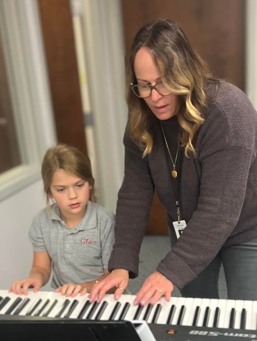 Woman teaching a young girl to play piano, indoors. The woman points at keys, girl watches.