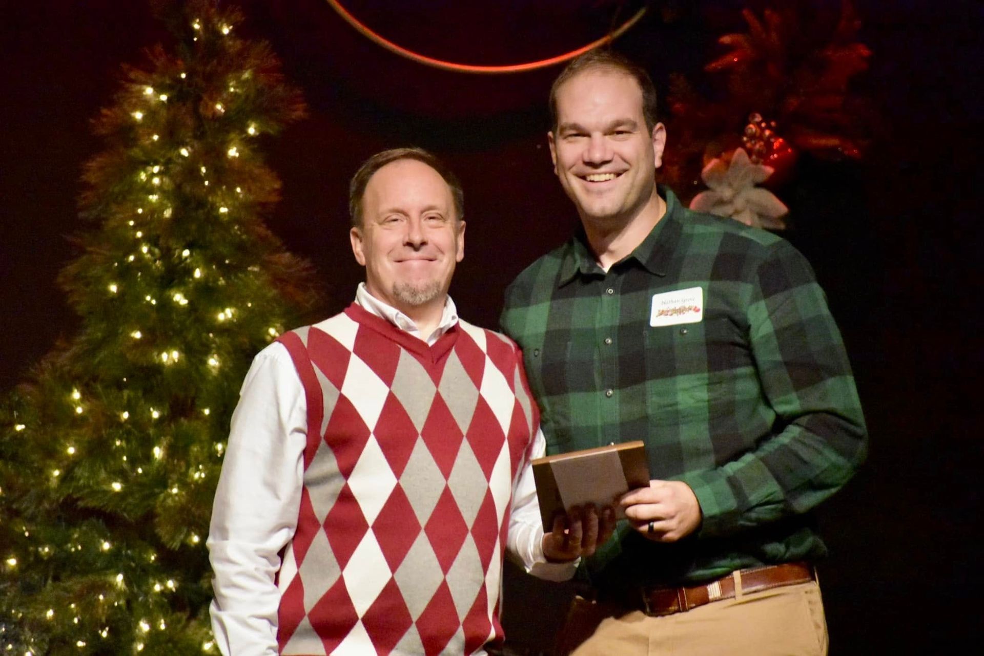 Two men standing by a Christmas tree. One holds a trophy, both smile. Man on left wears argyle sweater. Man on right wears plaid.