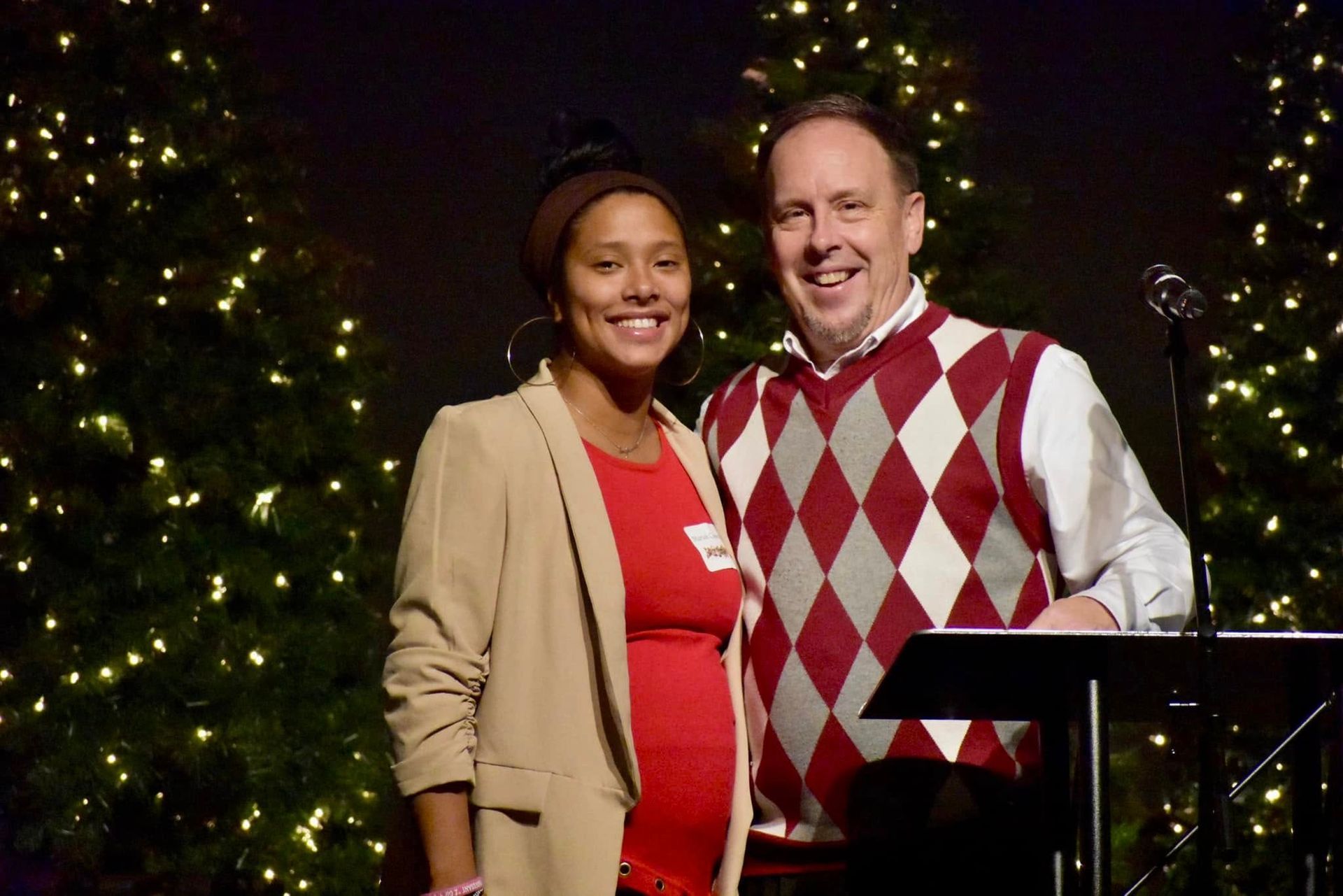 Woman and man smiling in front of lit Christmas trees. Woman in tan blazer, man in argyle sweater, at podium.