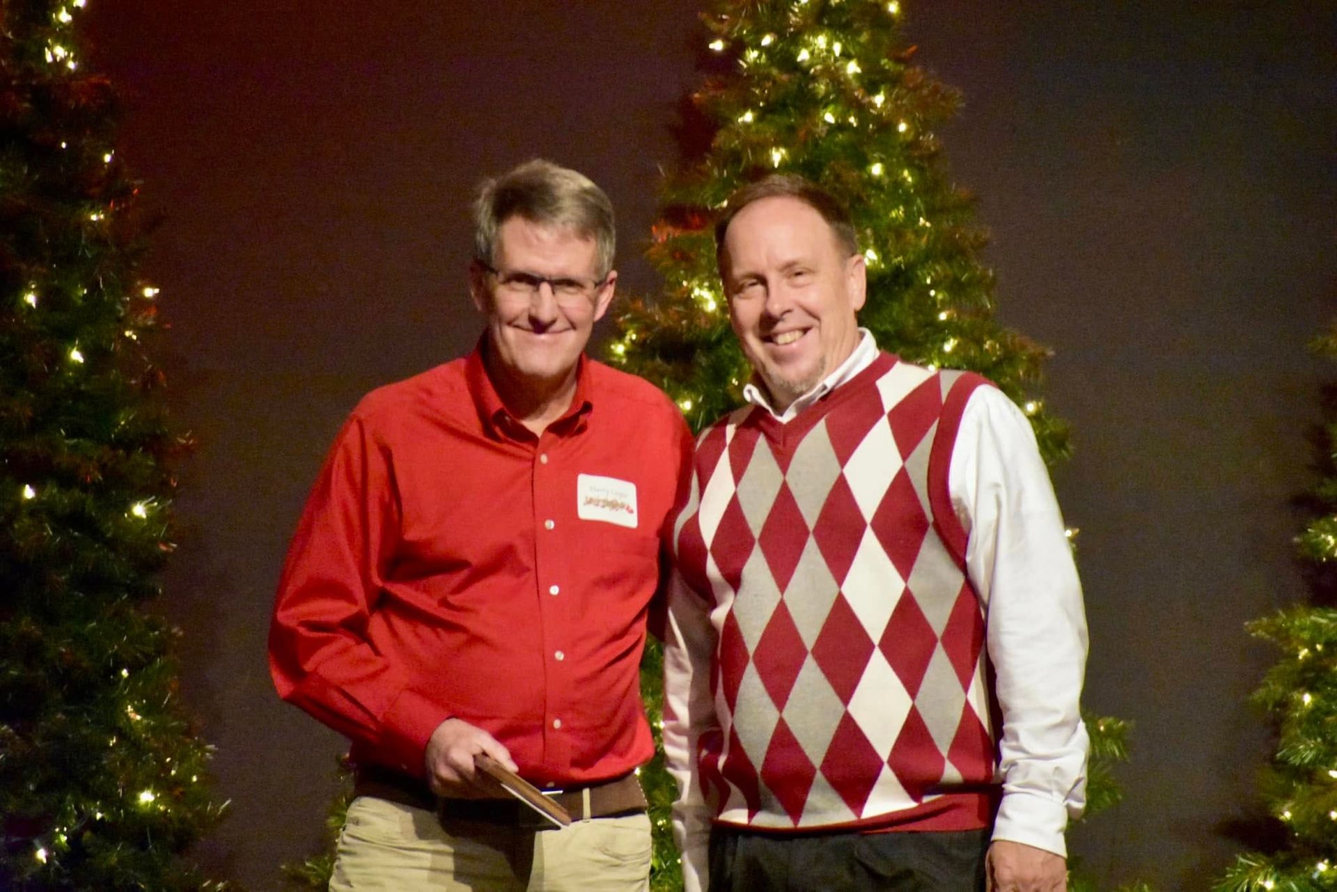 Two men smiling, posing in front of Christmas trees. One in red shirt, other in argyle sweater.