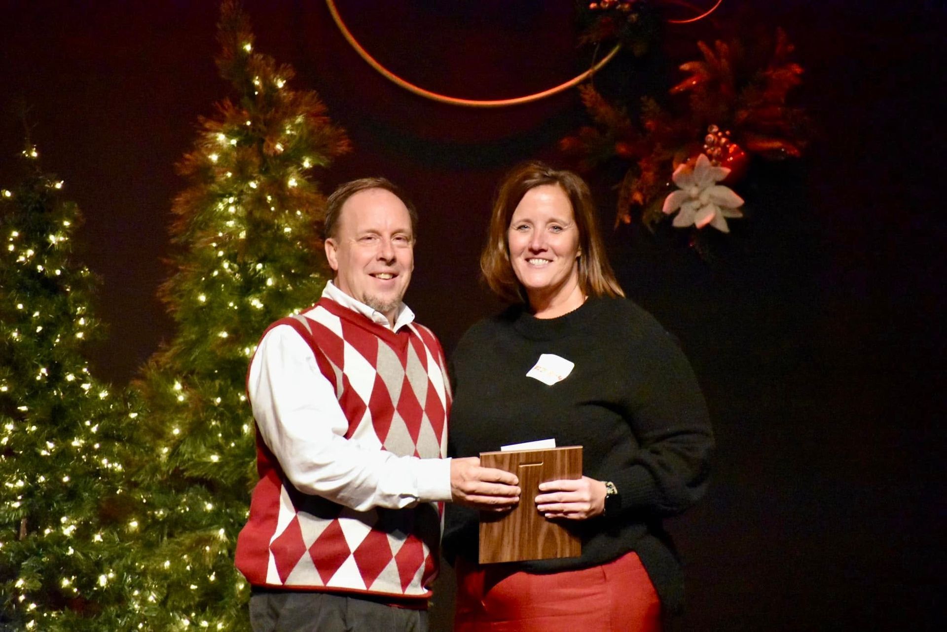 Man in argyle sweater presents a wooden plaque to a woman in a black shirt and red skirt; festive backdrop.