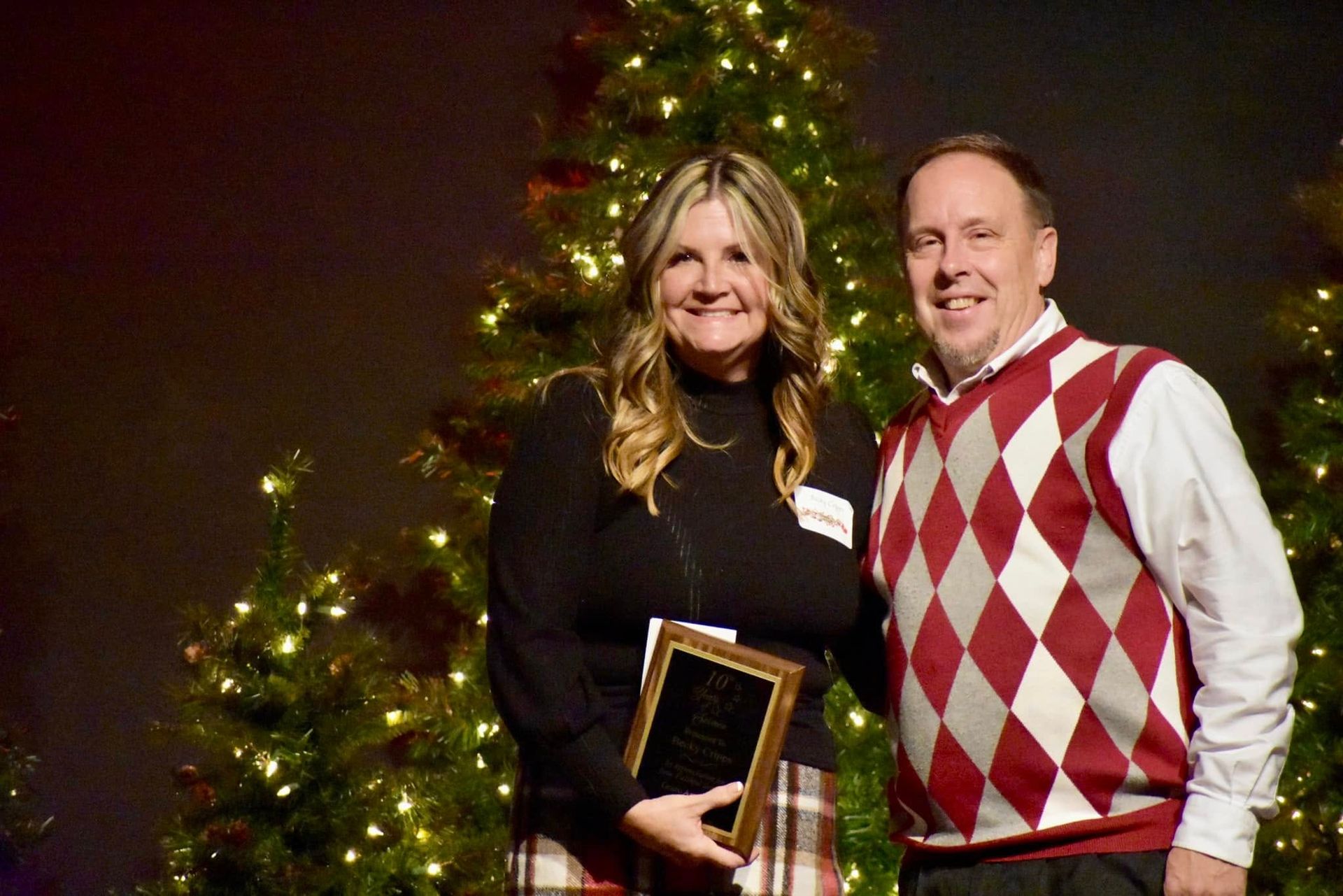 Woman holding award, smiles with man in front of Christmas trees, both looking at the camera.
