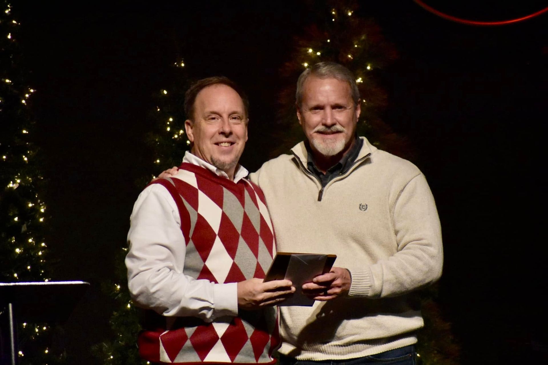 Two men stand together, one holding an award, both smiling. Background is dark with Christmas decorations.