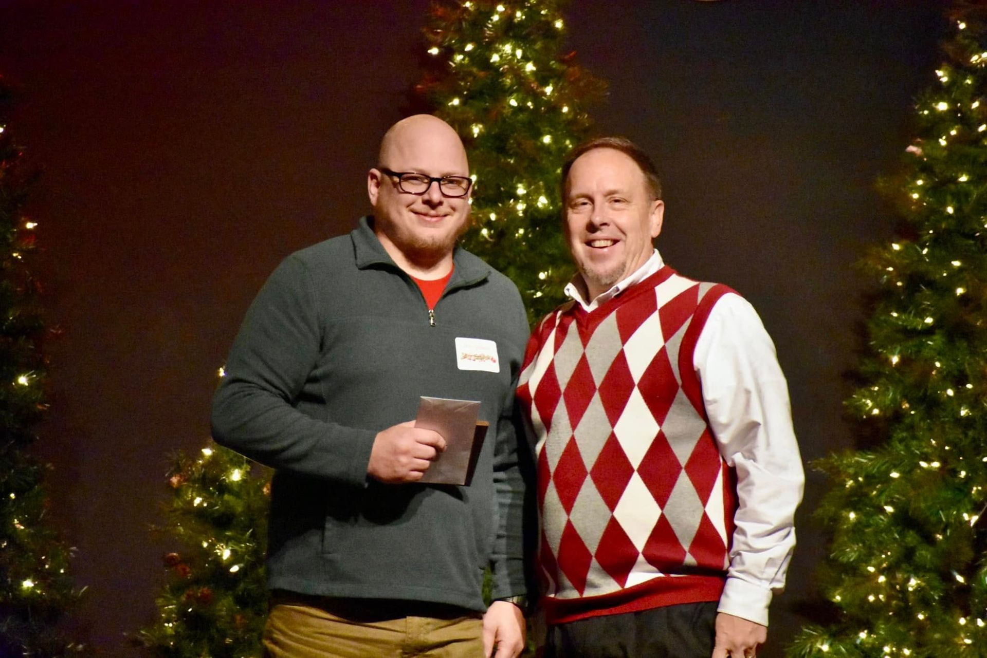 Two men posing in front of Christmas trees. One holds a paper, wearing glasses and a gray sweater. The other wears a red and white argyle sweater.