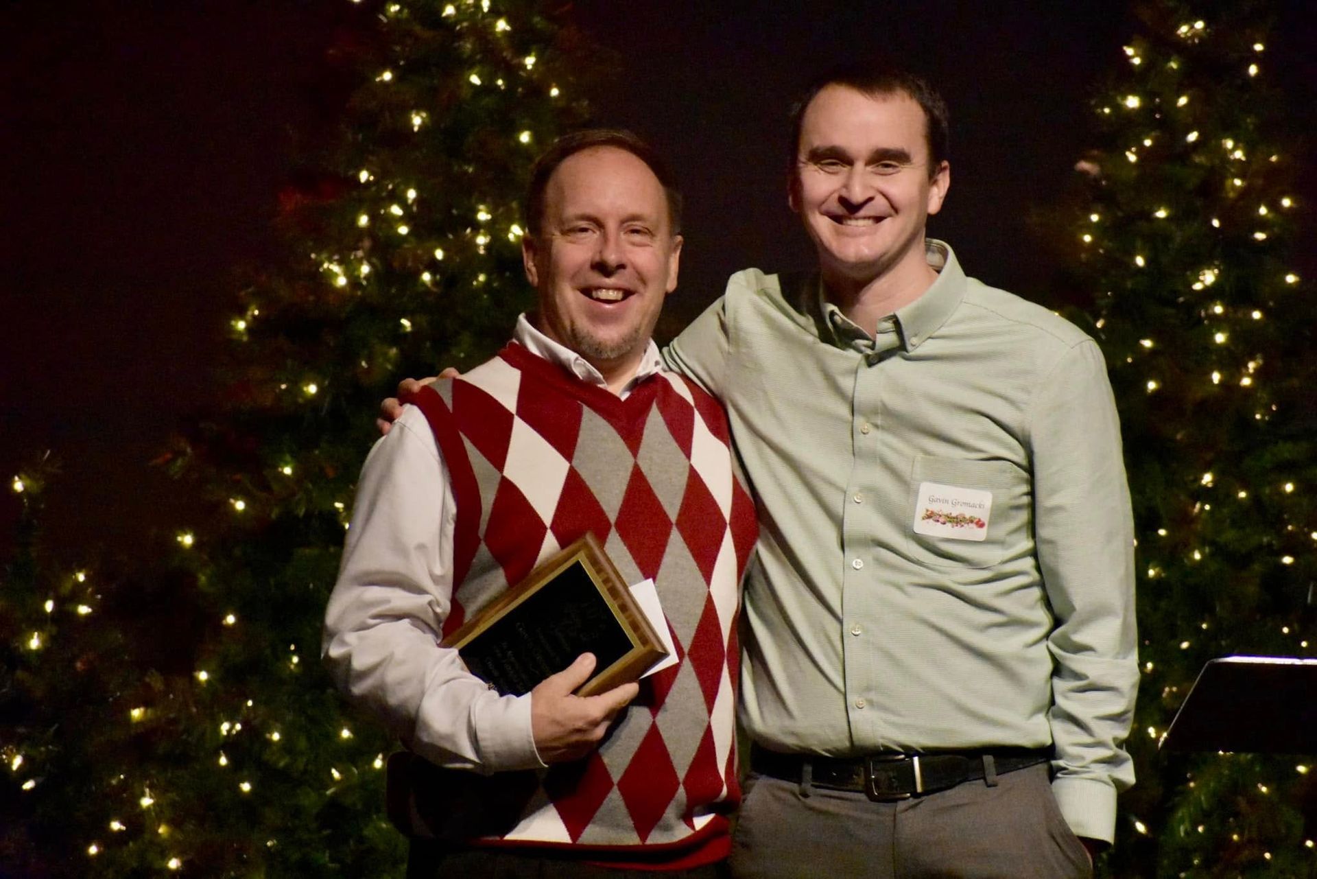 Two men smiling, posing by lit Christmas trees. One wears a argyle vest, holding a book; the other, a green shirt.