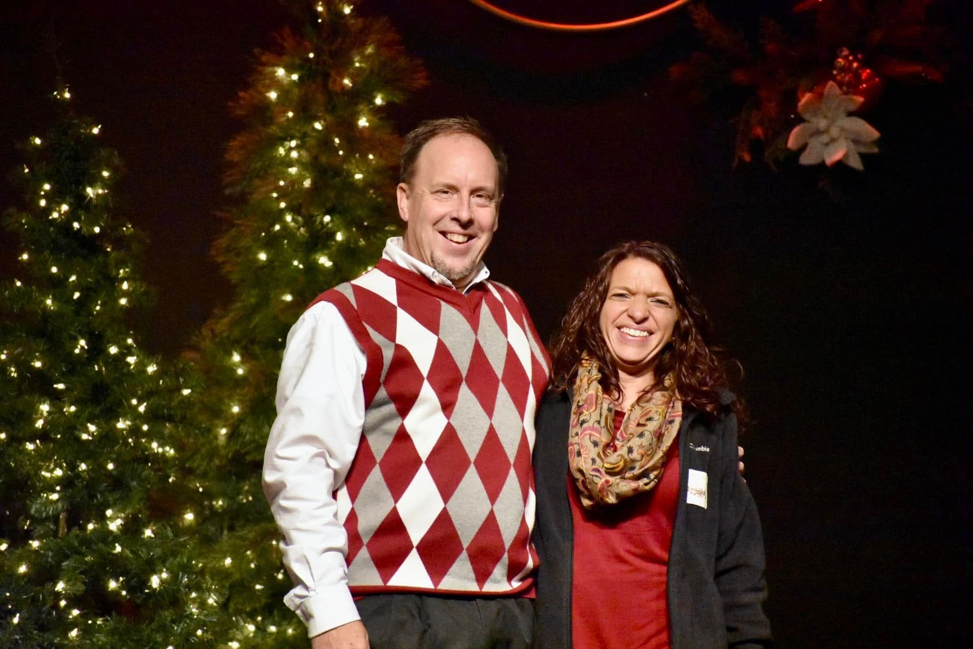 Man and woman smiling in front of a Christmas tree. Man in argyle sweater, woman in scarf. Dark background.