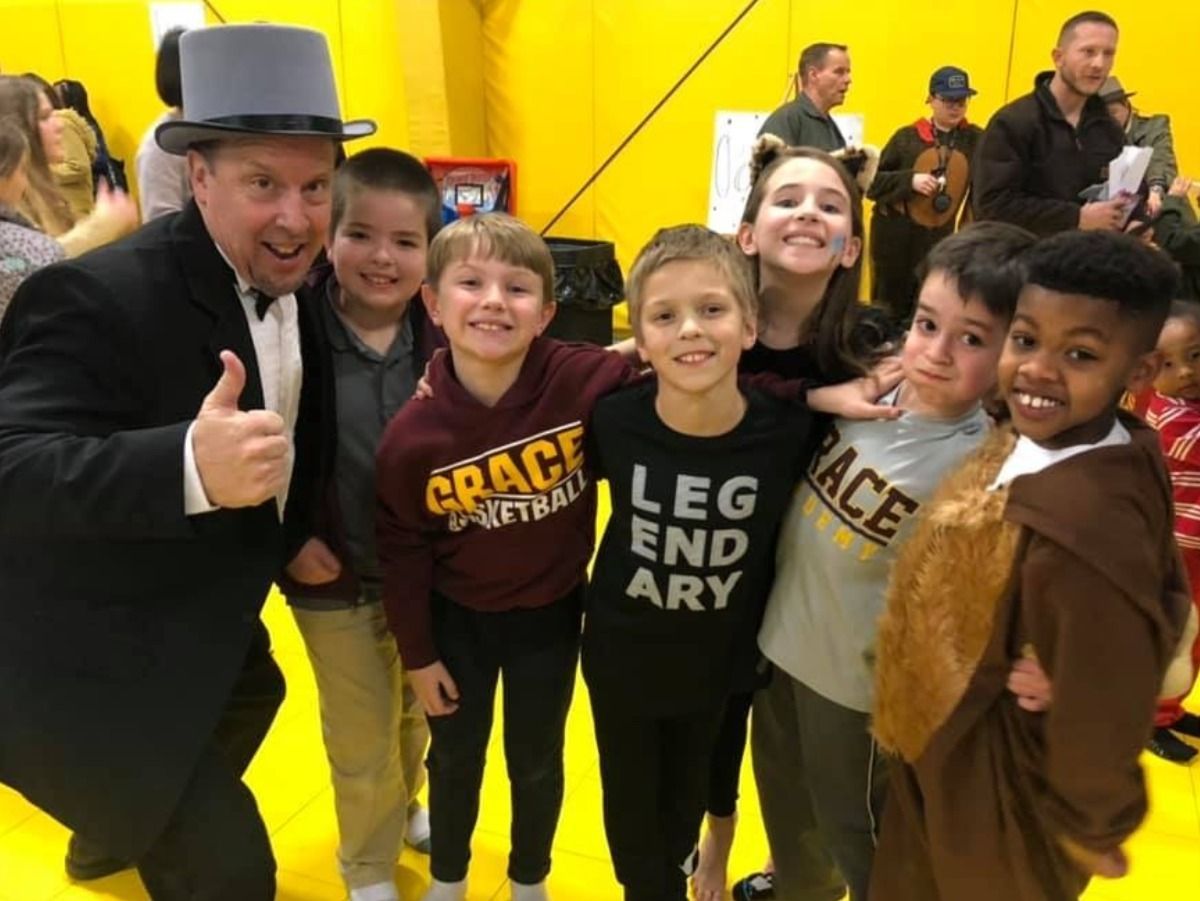Magician with kids, smiling, thumbs up, at event. Indoor, yellow wall. Kids in costumes and casual wear.