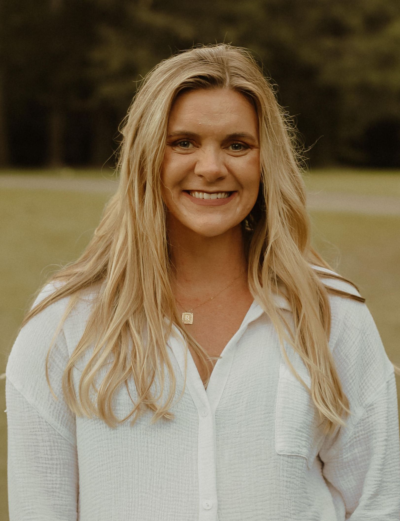 Woman with long blonde hair smiles, wearing a white button-down shirt outdoors.