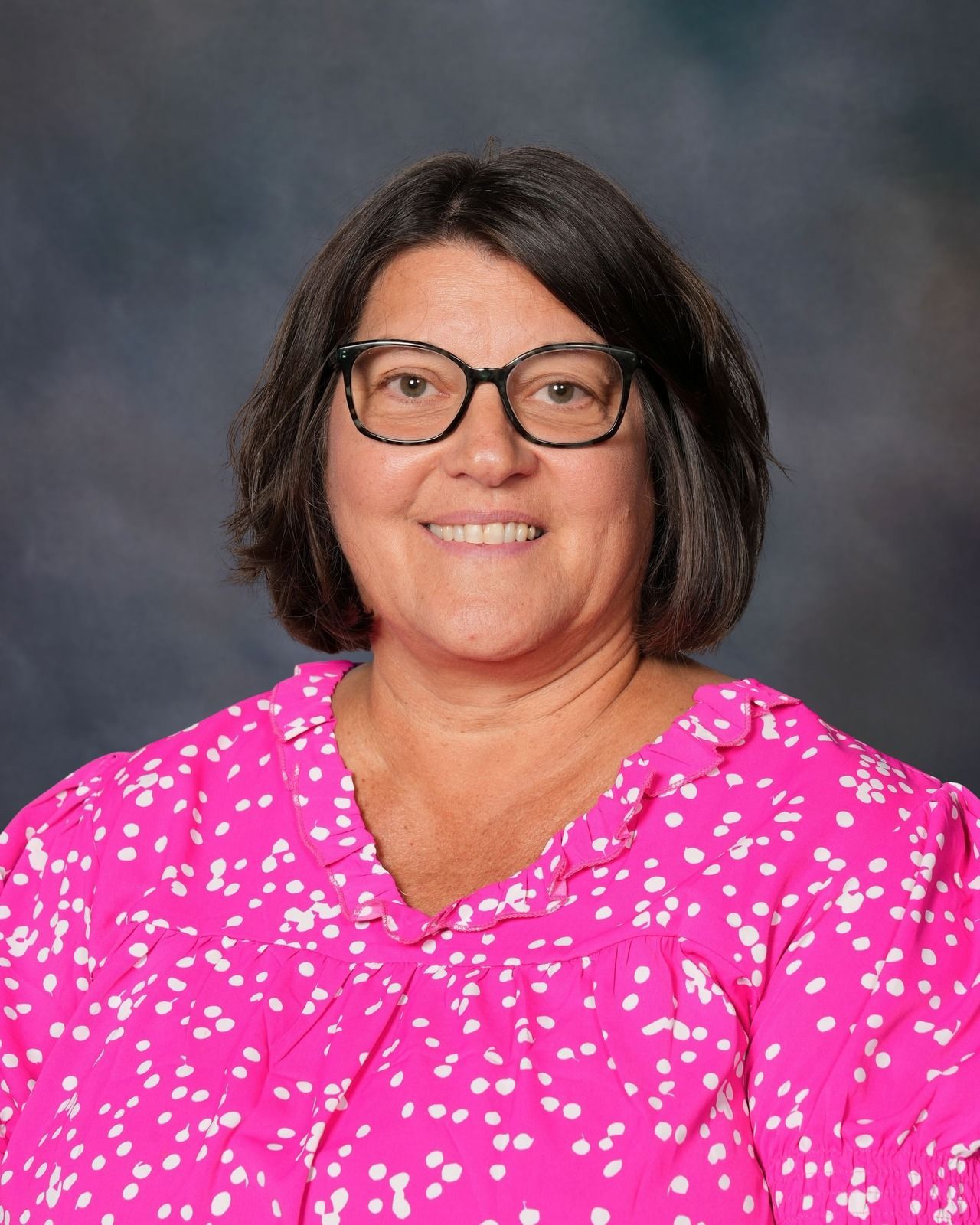 Woman with glasses smiling, wearing a blue floral shirt, gray backdrop.
