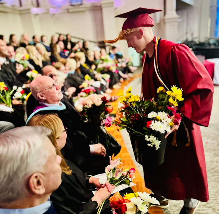 Graduate in maroon gown offers flowers to someone in the audience at graduation.