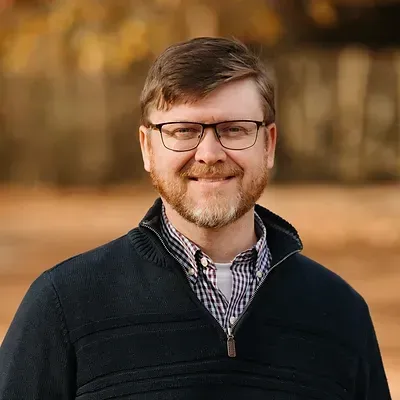 Man with glasses and beard smiles outdoors, wearing blue sweater over checkered shirt.