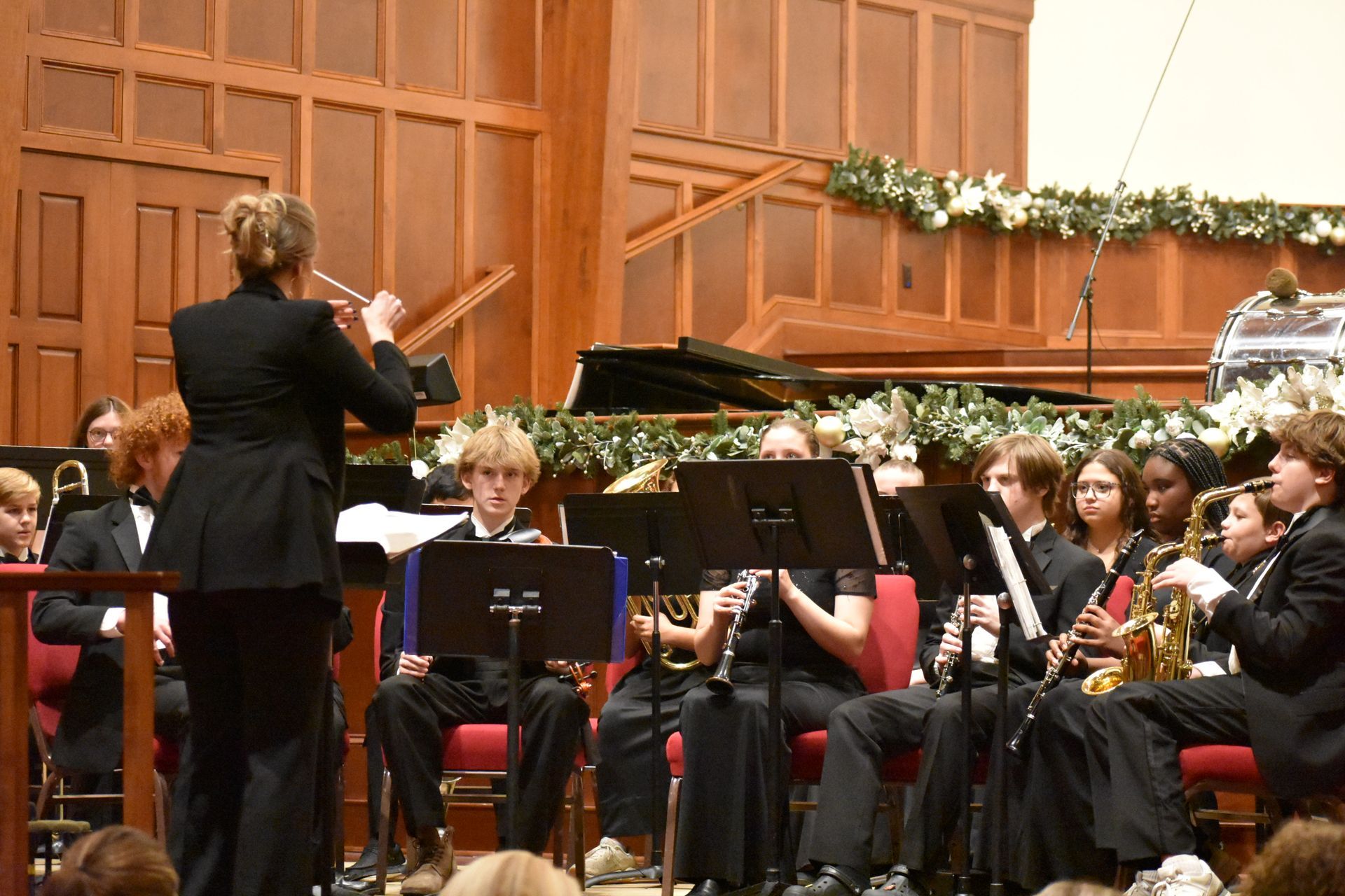 A band conductor leads a youth orchestra in a wood-paneled concert hall.