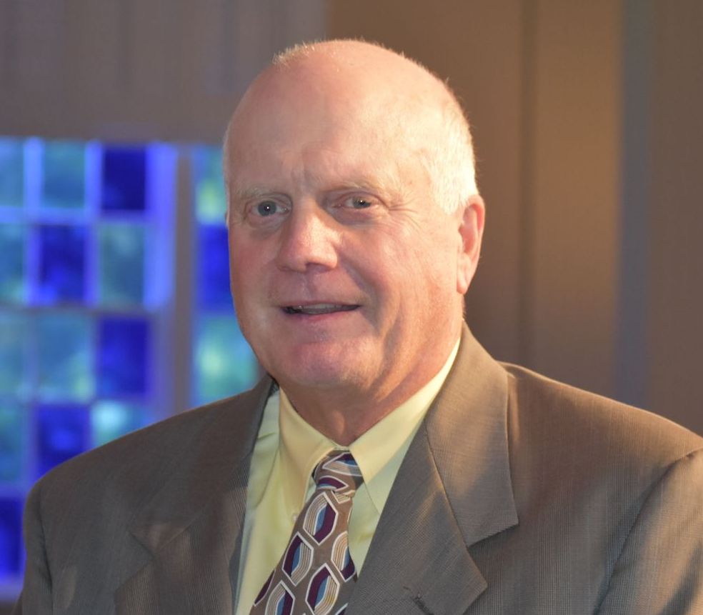 Bald man in a suit, smiling; background with blue and green glass blocks.