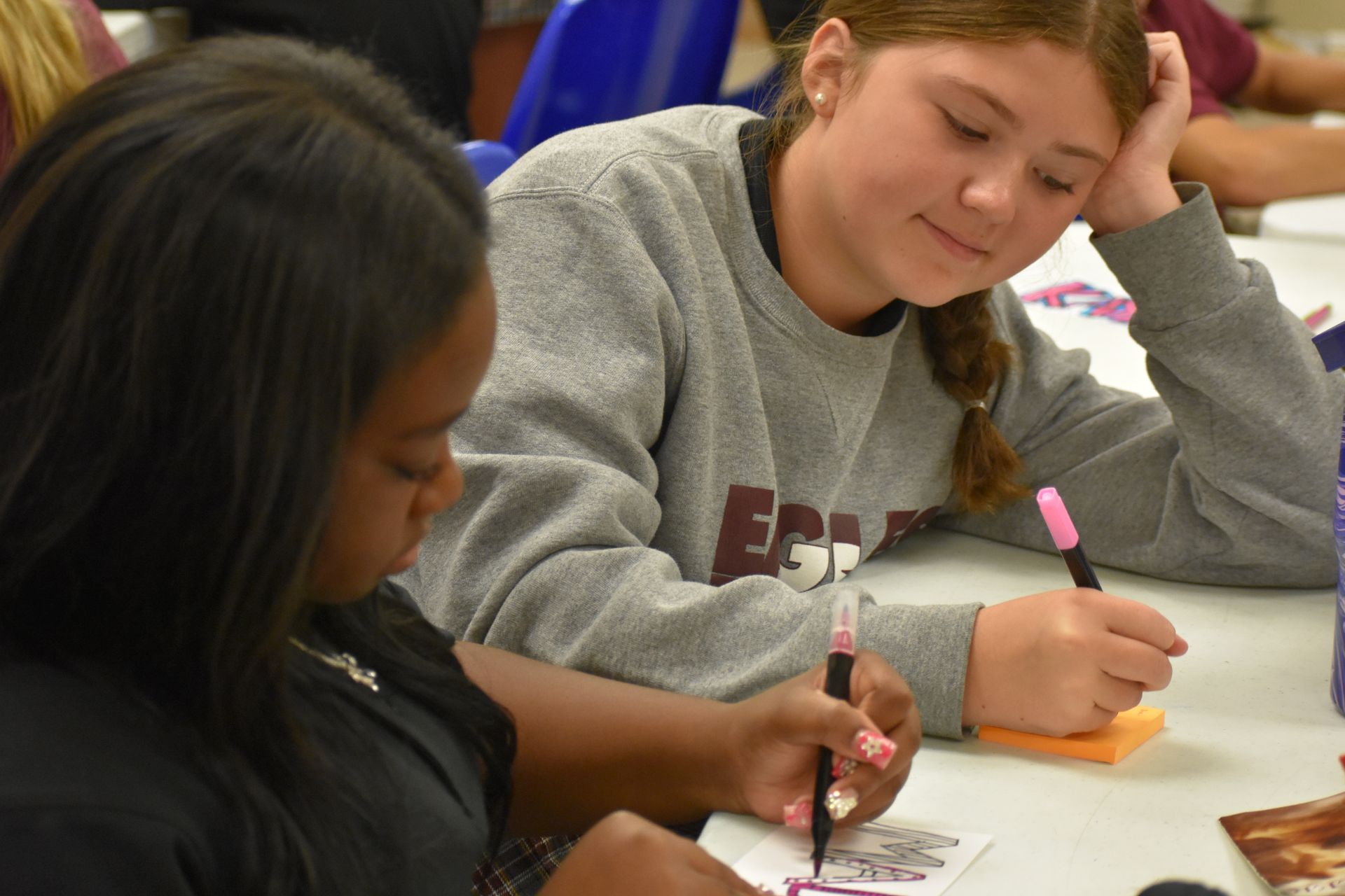 Two young women, one with dark hair, one with a braid, writing with pens at a table.