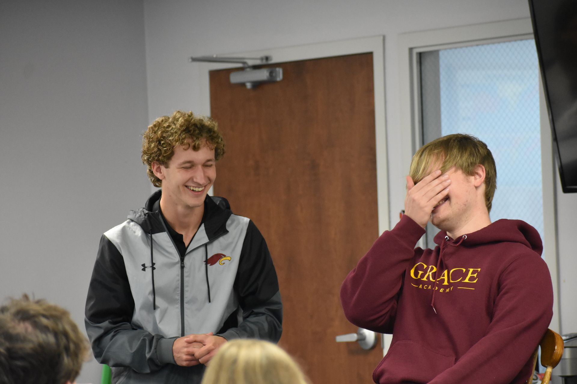 Two young men laugh; one covers his face. Indoors, possibly a classroom.