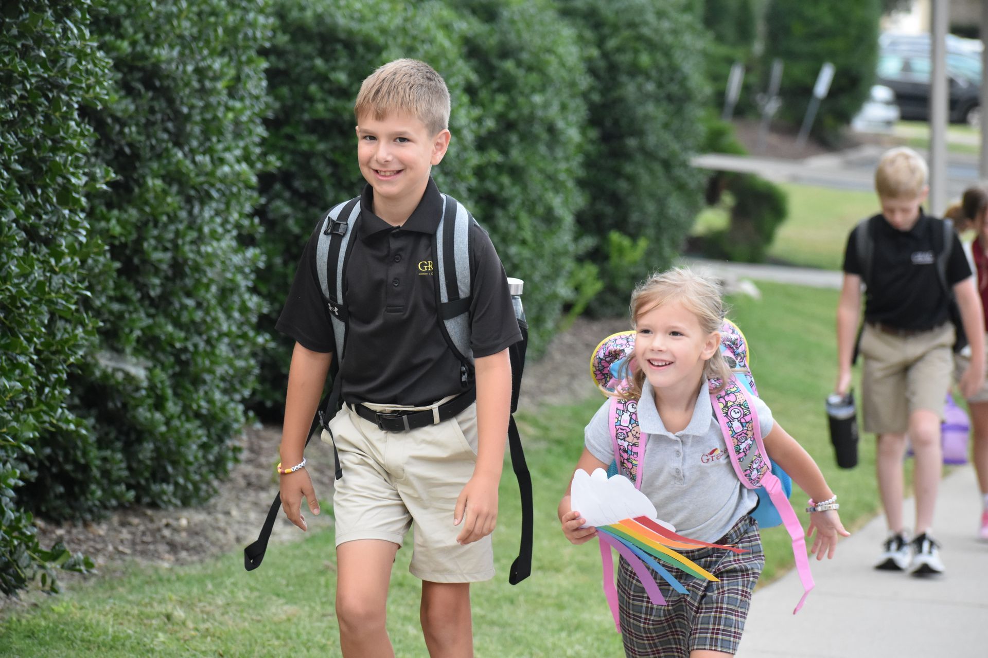 Two students, smiling, walking on a sidewalk next to a hedge. Boy in black shirt and khaki shorts. Girl holding paper rainbow.