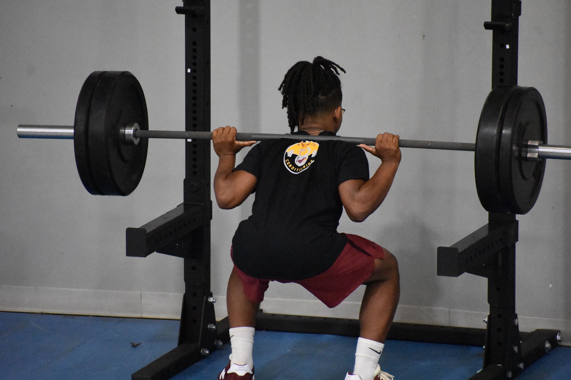 A young person with dreadlocks squats with a barbell in a gym, weight lifting.