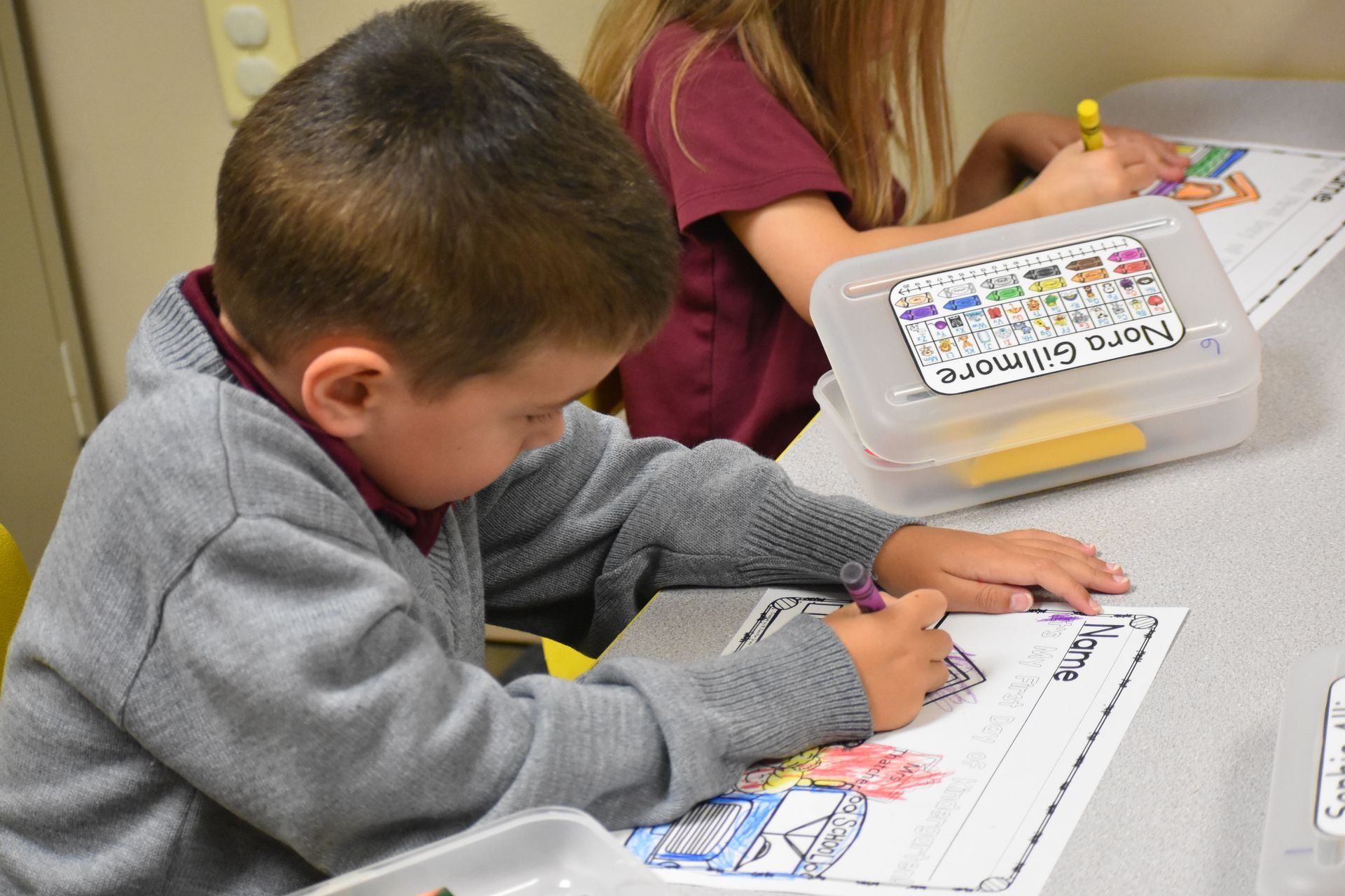 Boy coloring at a desk, using a purple marker. Another child nearby; white plastic box labeled 