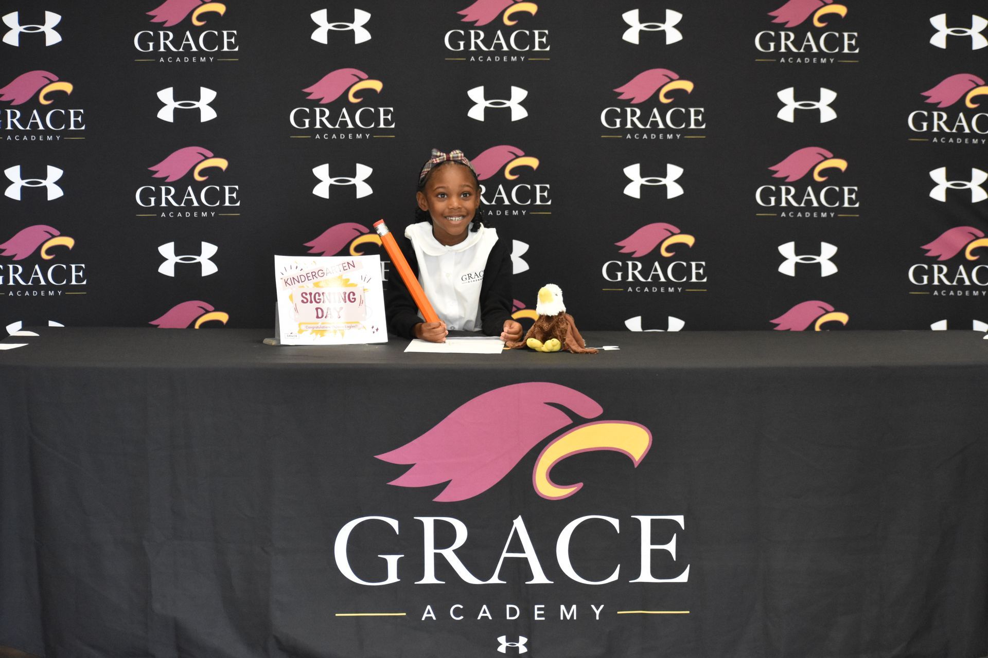 Young girl at a Grace Academy signing table, smiling, drawing. Backdrop has school logo and sponsor logos.