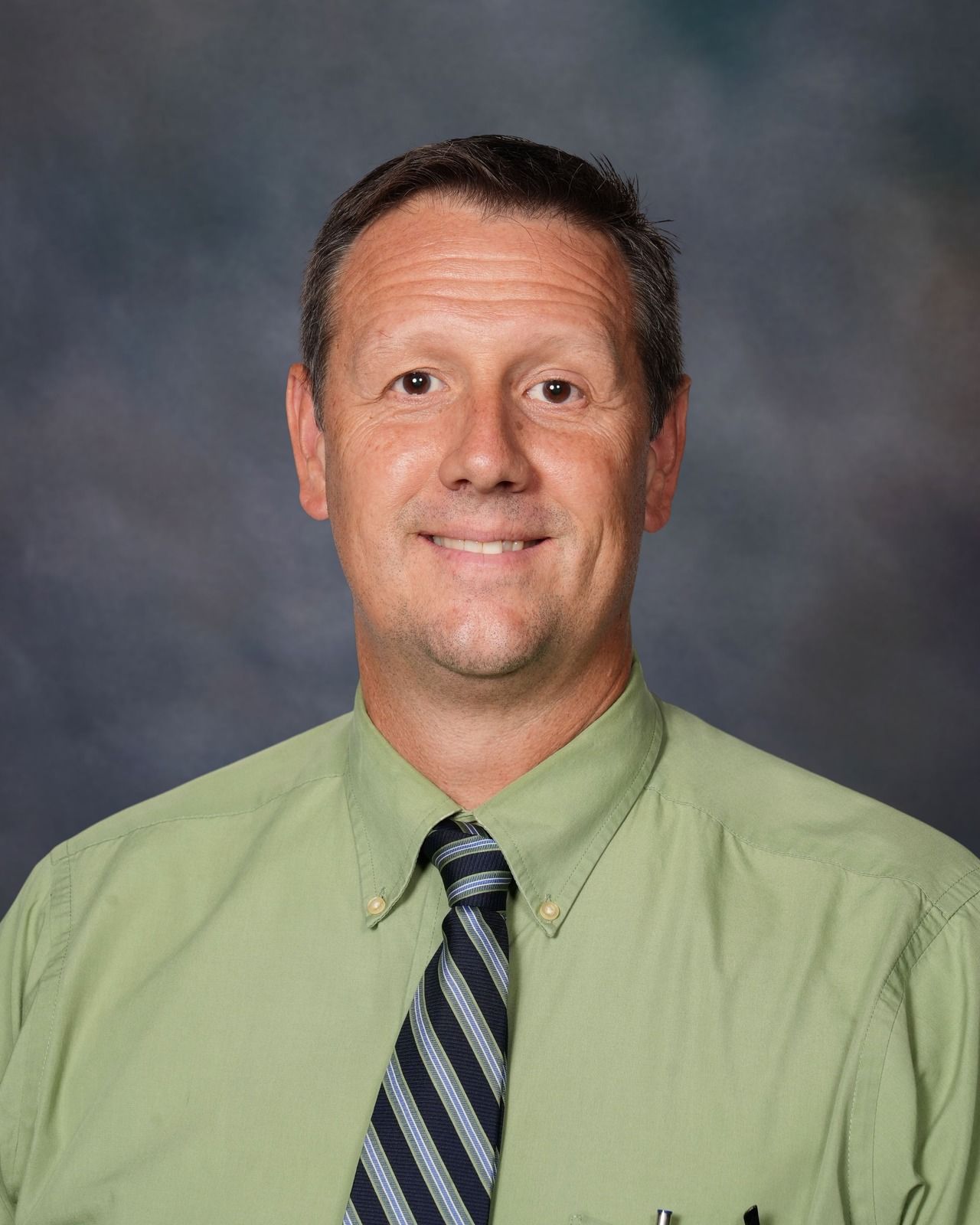 Man with brown hair wearing a white shirt and patterned tie, smiling at the camera, against a gray backdrop.