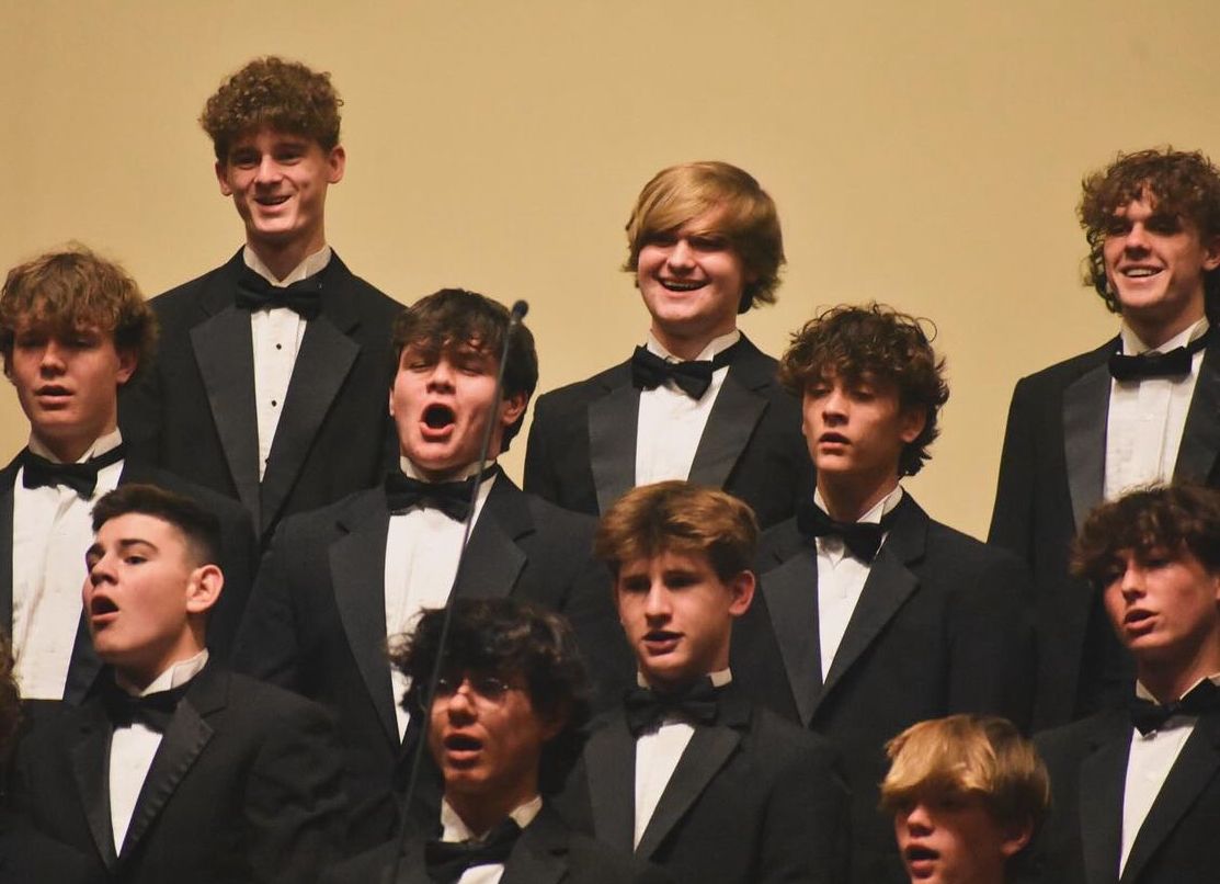 Male choir in formal wear singing on stage; neutral background.