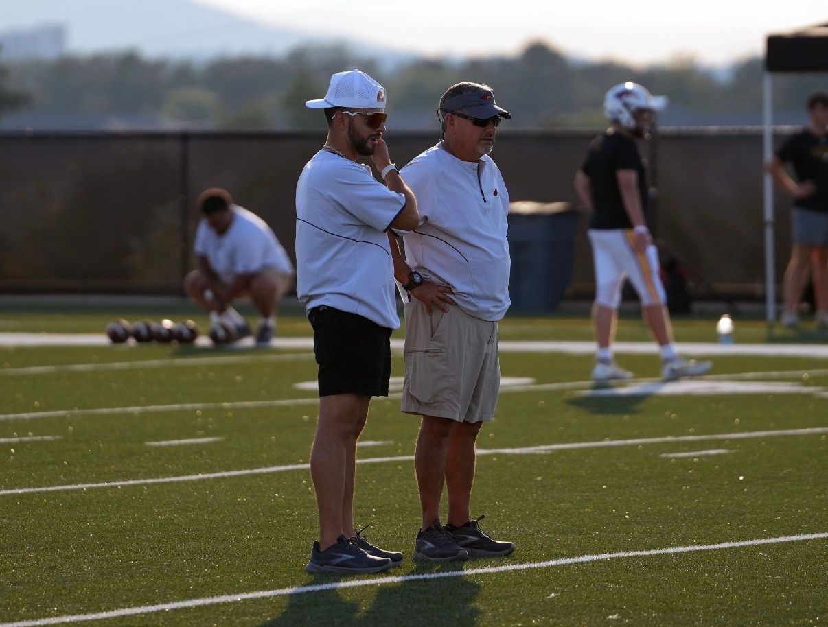 Three men in maroon shirts and shorts/pants stand on a sports field, looking down with closed eyes.