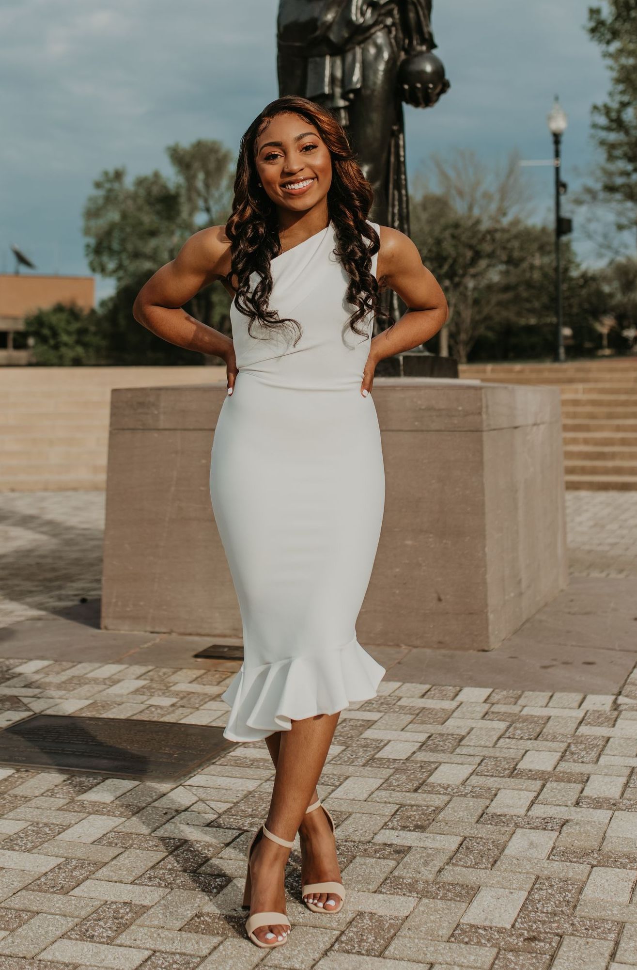 Woman in white dress poses smiling outdoors near a statue and brick walkway.