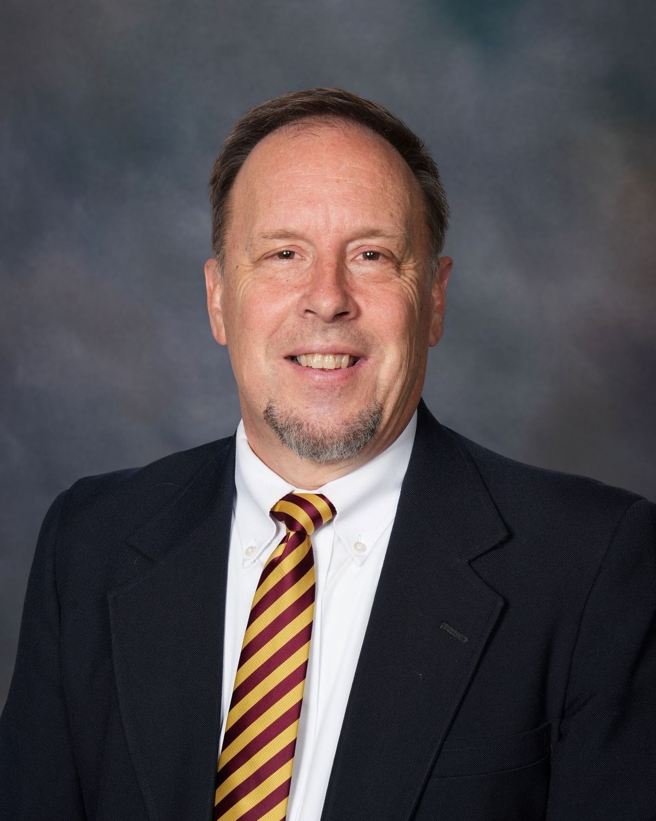 Man in blue suit, smiling, wearing a striped gold and maroon bow tie.