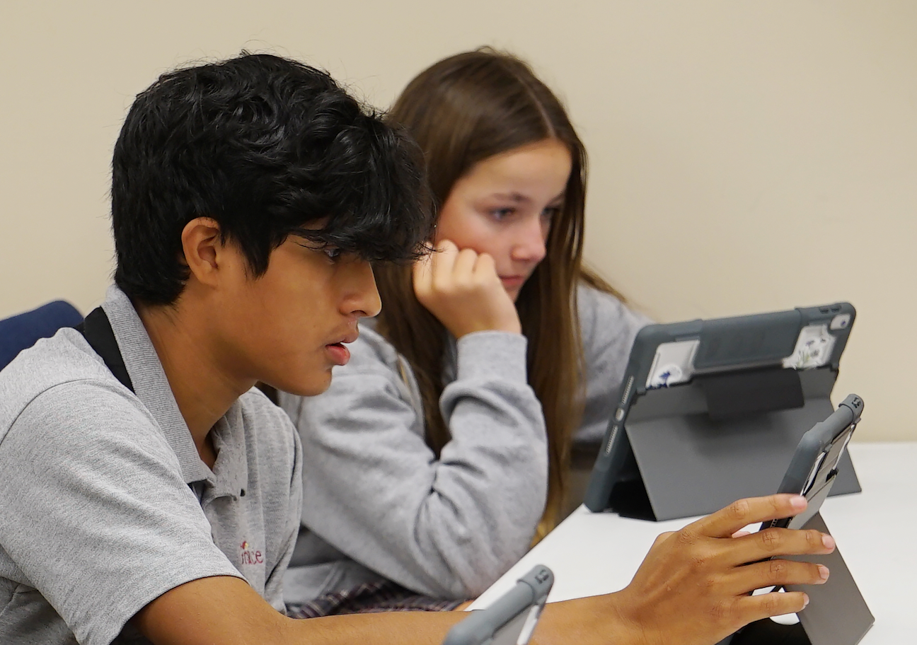 Two students look at tablets at a table; boy points, girl watches; indoors, light colors.