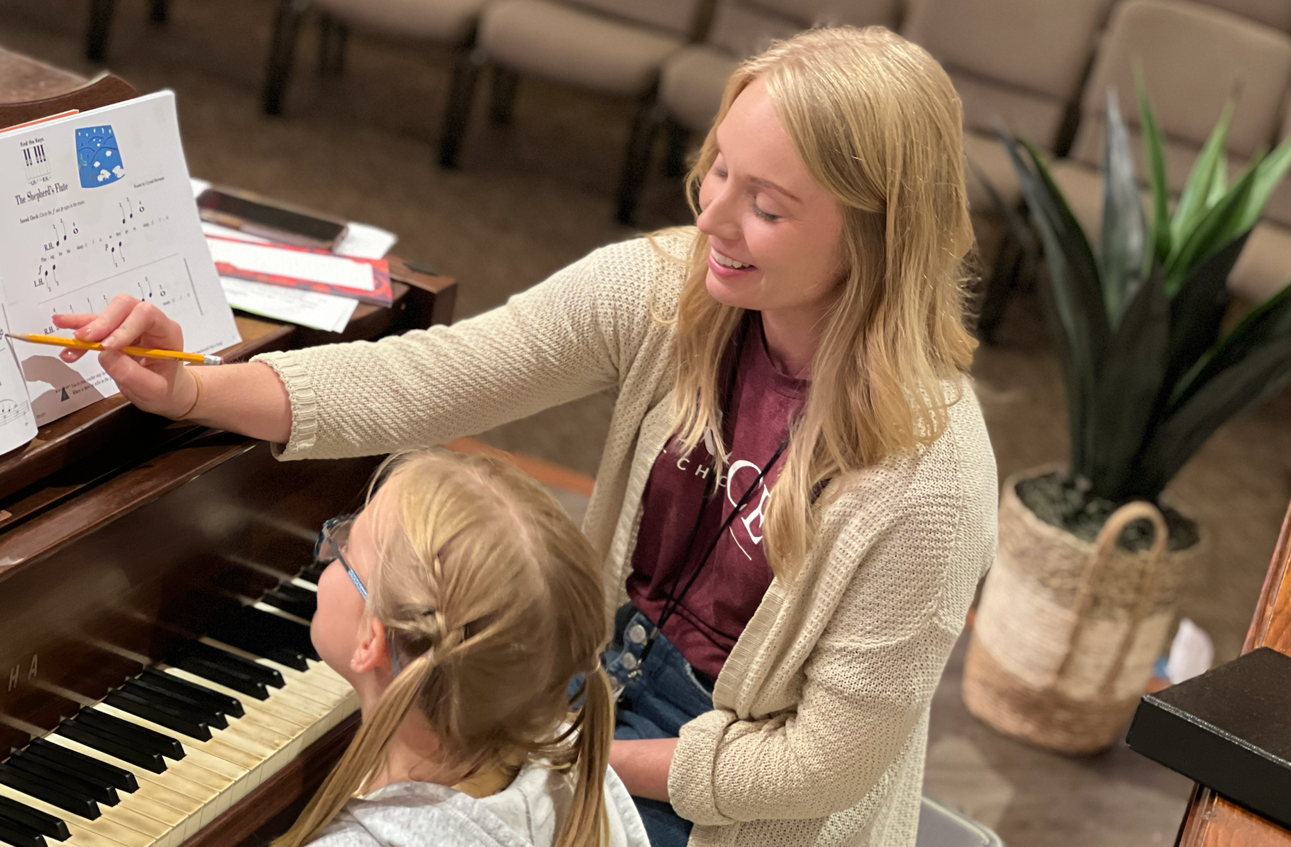 Woman with blonde hair teaches a young girl piano, pointing at sheet music. Inside, natural light.