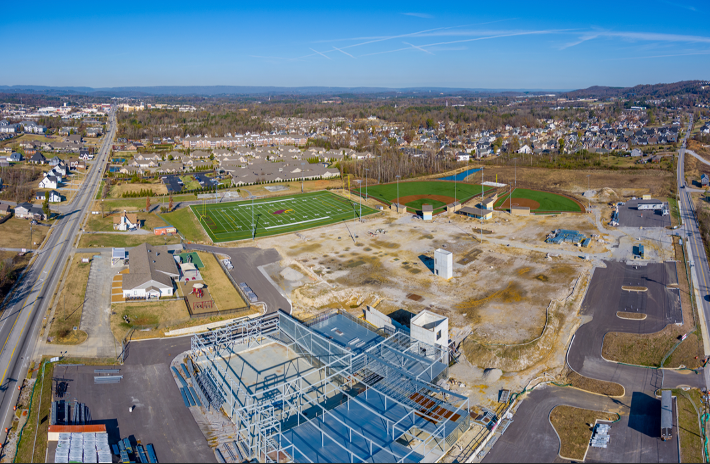 Aerial view: Construction site with metal framing, sports field, and town in the background.