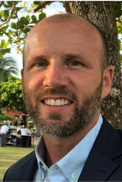 Man with beard smiling, wearing a suit jacket, outdoors near a tree.