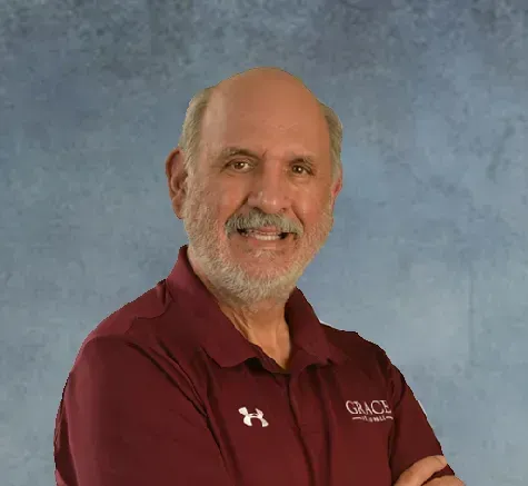 Smiling bald man with a gray beard wearing a maroon polo shirt with crossed arms.