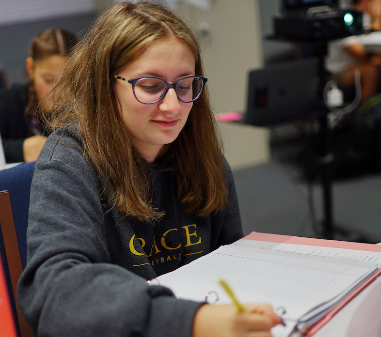 A young woman with glasses writes in a notebook, smiling. She wears a gray sweatshirt, indoors.