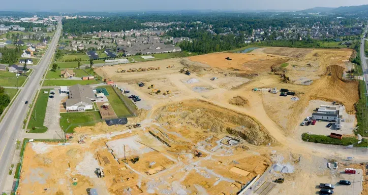 Aerial view of a large construction site with exposed earth, machinery, and nearby buildings on a sunny day.