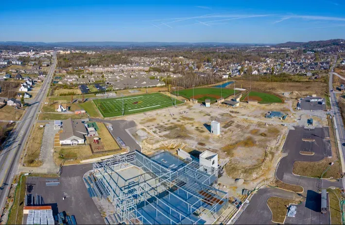 Aerial view of a construction site, sports fields, and a town. Blue steel frame of a building under construction.