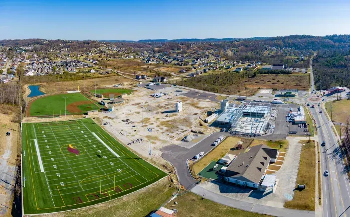 Aerial view: football field, construction site, road, and distant houses under a clear sky.