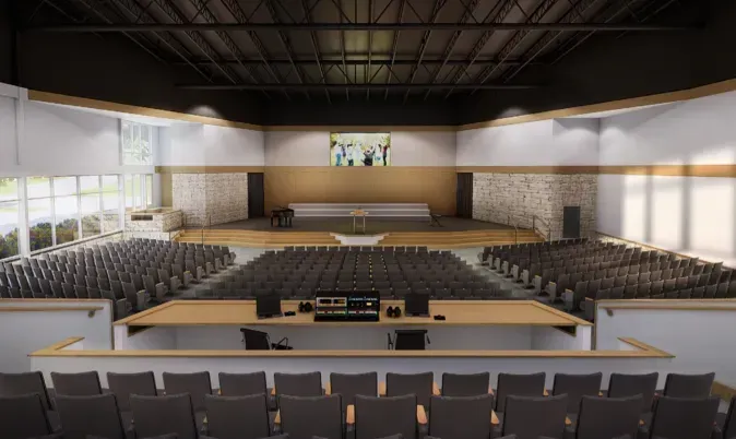 Auditorium with rows of seats, stage, soundboard, and video screen, inside a modern building with a dark ceiling and white walls.