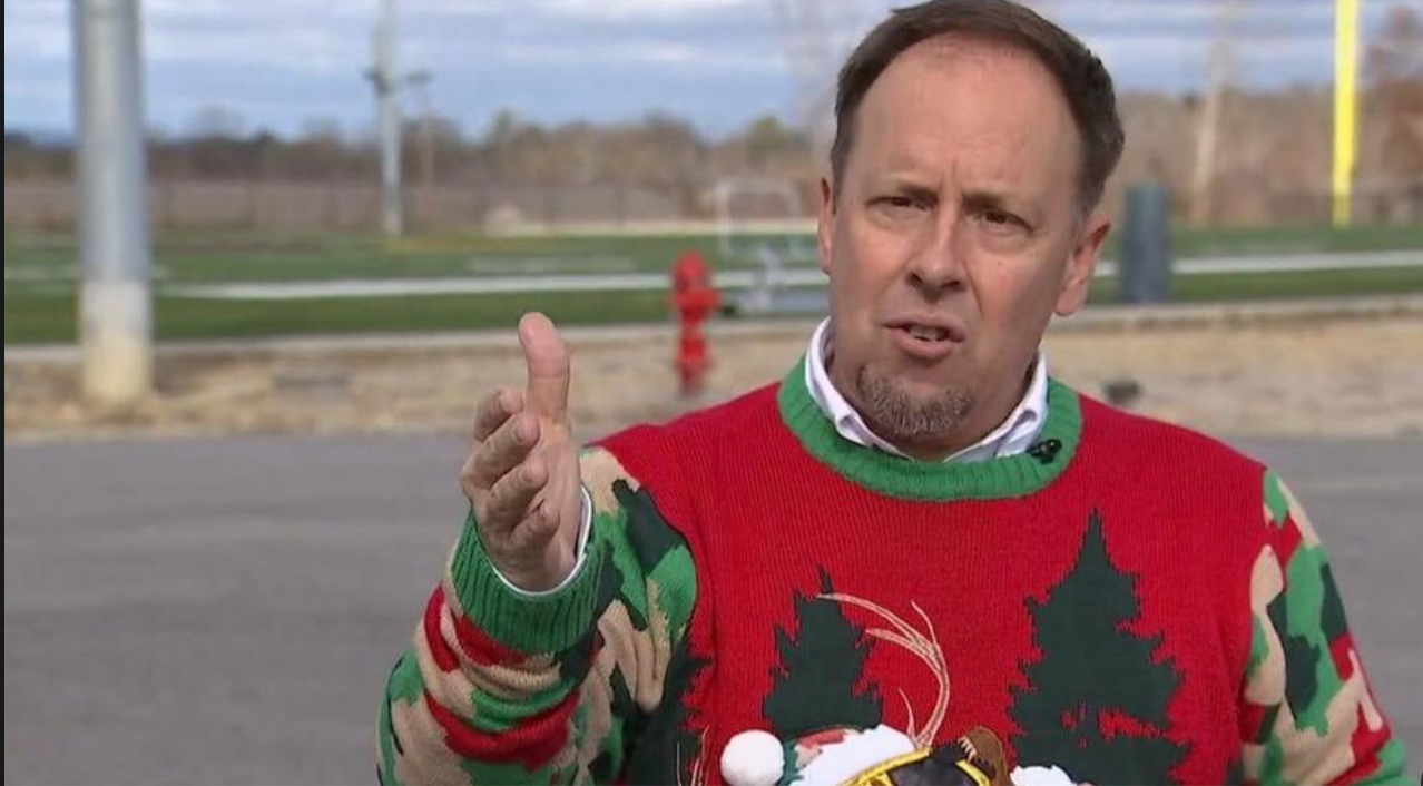 Man in Christmas sweater gesturing outside; trees, and field in background.