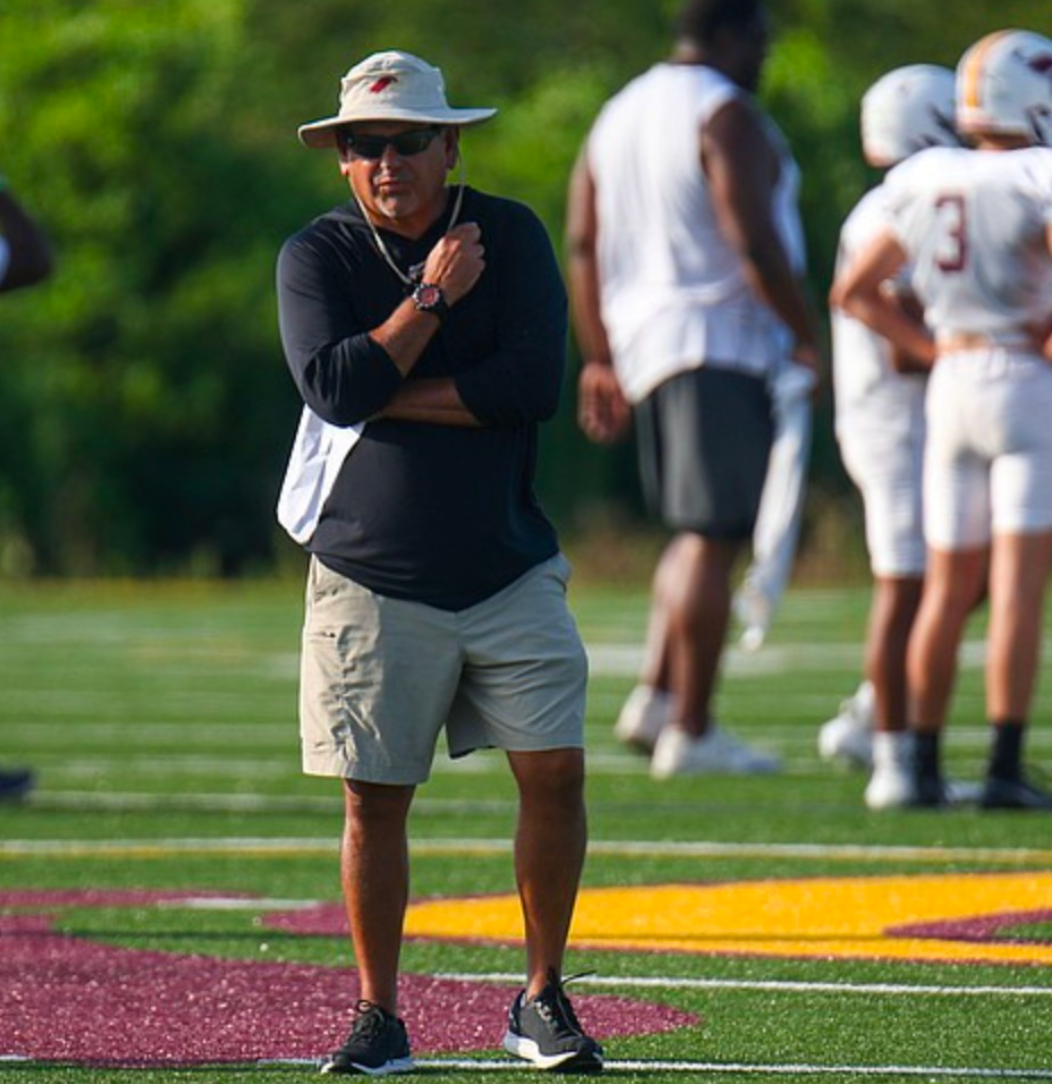 Man in a sun hat, khaki shorts, and a black shirt watches football players on a green field.