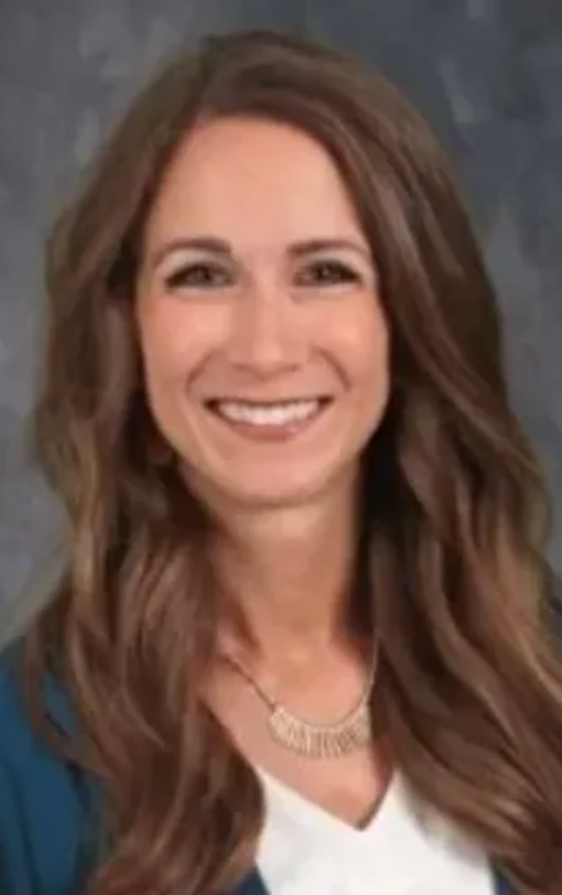 Woman with long brown hair smiles, wearing a blue blazer and necklace.